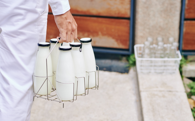 A milkman's arm holding a 6-pack of milk bottles as he walks towards a front door.